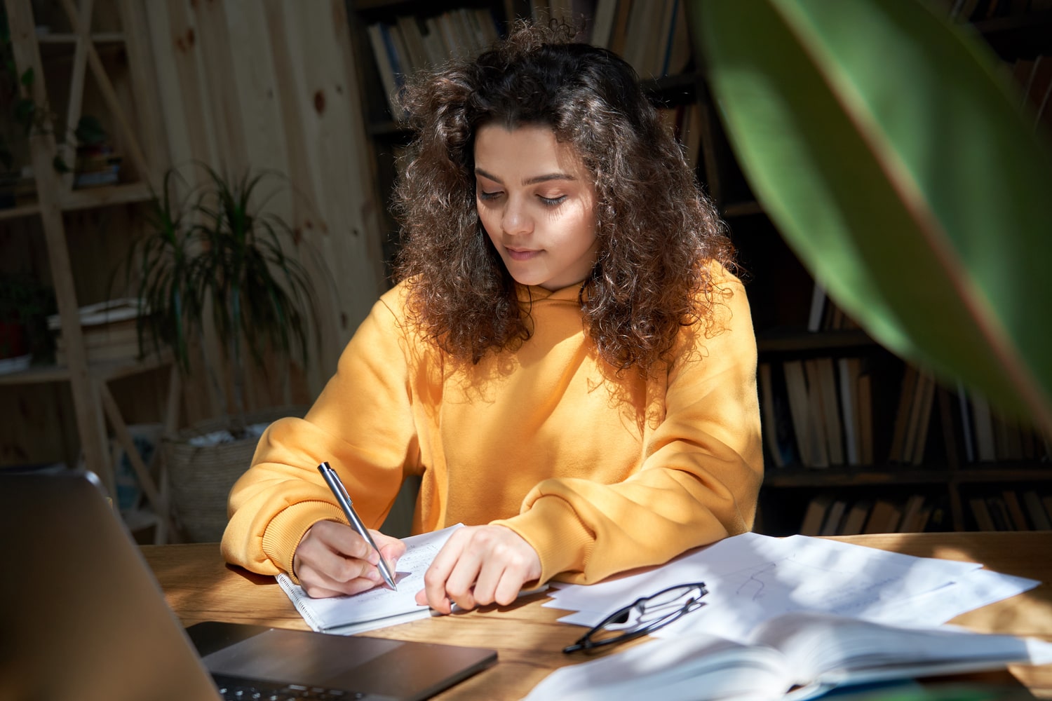 girl in yellow sweatshirt sitting at a table with her notebook and laptop writing an essay